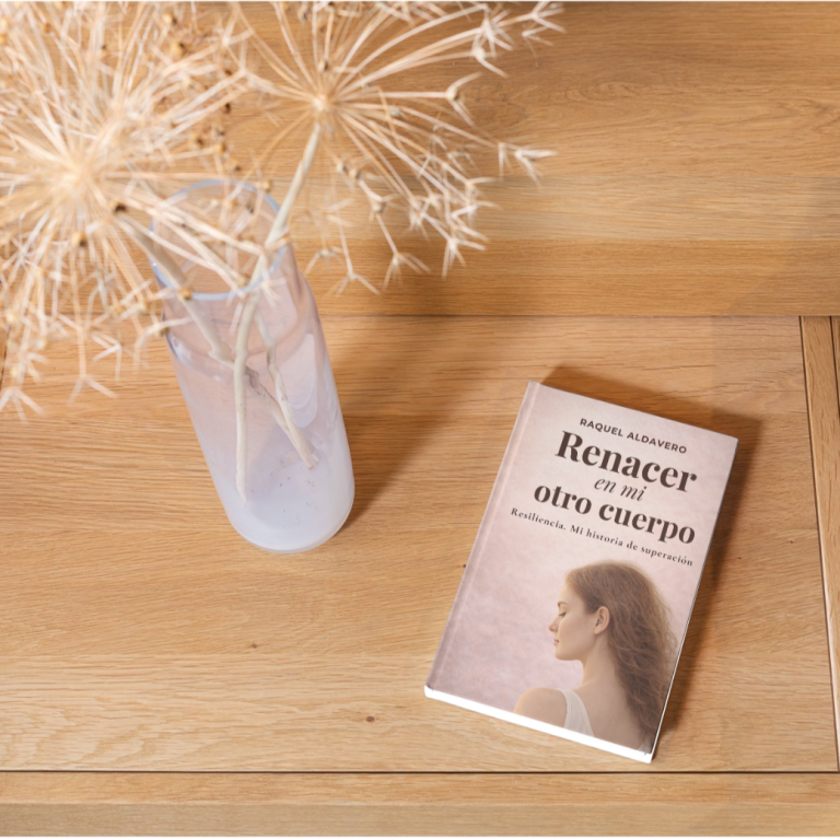 Un libro titulado "Renacer en mi otro cuerpo " junto a una flor seca en un jarrón.