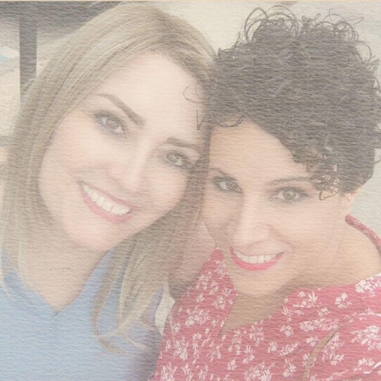 Dos mujeres sonrientes posando juntas al aire libre. Una lleva blusa azul y la otra roja.