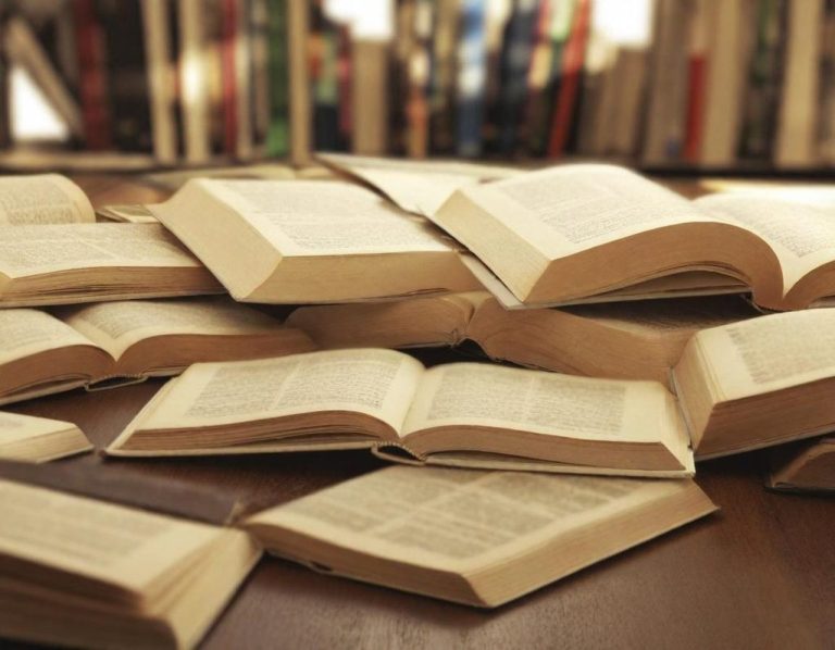 Libros abiertos desordenados sobre una mesa, con estanterías de fondo.